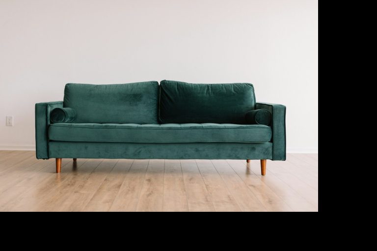Grüner Samtsofa mit Holzbeinen vor einer weißen Wand, minimalistisch und modern.