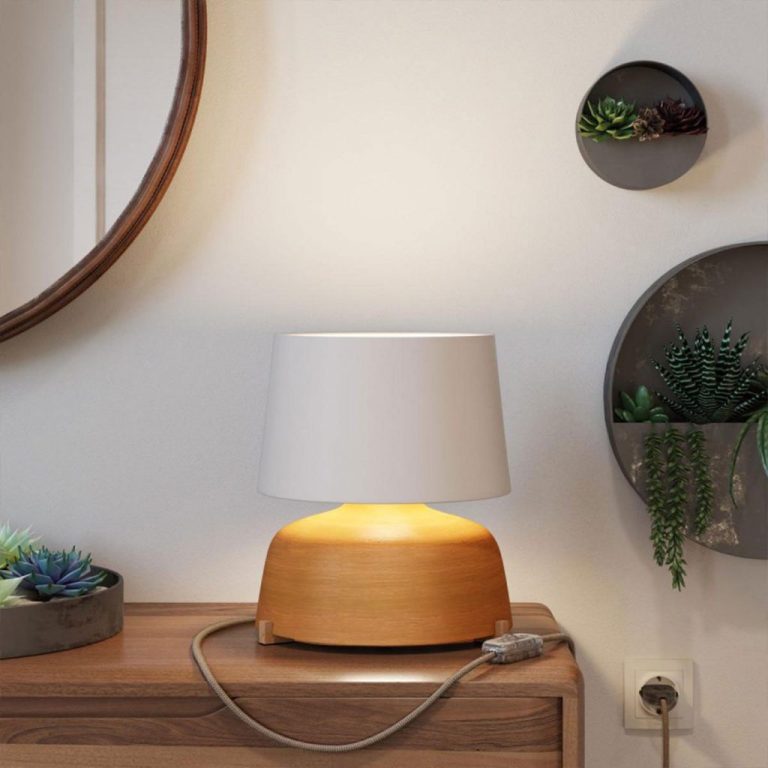 Holzlampe mit weißem Lampenschirm auf einem Holztisch, im Hintergrund Pflanzen.