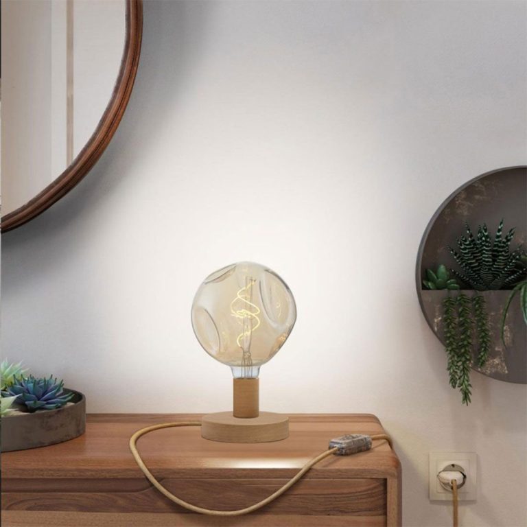 Tischlampe mit runder Glühbirne auf einem Holzsideboard vor einer Wand. Pflanze im Hintergrund.