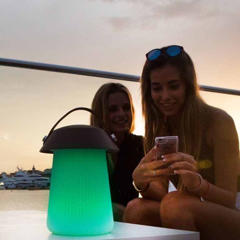 Zwei Frauen sitzen bei Sonnenuntergang und schauen auf ein Handy. Neben ihnen leuchtet eine Lampe.